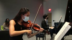 Video still of a woman in a face mask playing violin with a man in the background playing piano