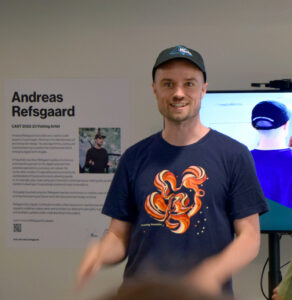 Andreas Refsgaard stands in front of a computer monitor in a t-shirt and baseball cap