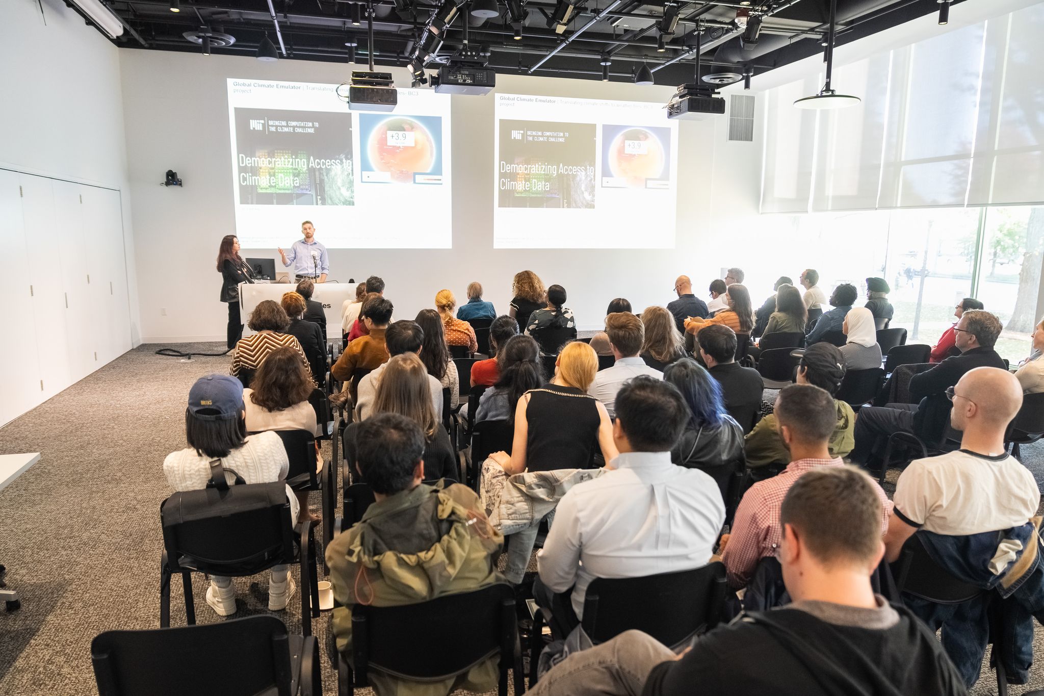 A seated audience watches a man and a woman standing at a podium; a slide on the wall behind them reads, 'Democratizing Access to Climate Data.'