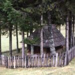 Color photograph of a small wooden building with a steeply pitched wooden hip roof surrounded by a ring of fir trees and a wooden picket fence on an open hillside.