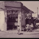 A photograph of a street in Baghdad, Iraq taken in 1976 that shows a corner shop with musical instruments such as trumpets and baritones hanging from the entrance.