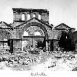 A black and white photograph of a ruined stone church in rural Syria, showing the triple-arched entryway. Many large stones dot the ground in front of the arch. The word “Entrée” is written in the blank space at the bottom of the image.