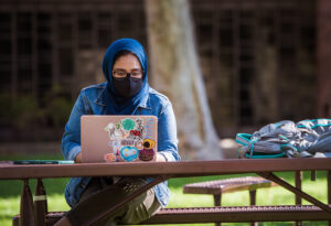 A woman in a head scarf and face mask works on a laptop at a picnic table outdoors