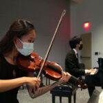 Video still of a woman in a face mask playing violin with a man in the background playing piano