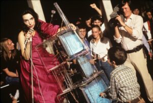 A woman in a red dress holds a microphone and leans over a stack of TVs encased in plexiglass; a crowd is behind her, a man is shooting video, and a young boy stands in front of her