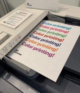A piece of paper sits in a printer; it reads "Color printing!" in several different colors