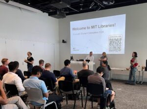 Three people stand at the front of a room before an audience; a slide on the screen behind them reads "Welcome to MIT Libraries! Graduate Student Orientation."