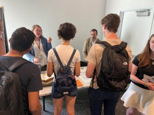 Student wearing backpacks stand in front of a table staffed by two people; there is a rare book on a cradle on the table.