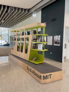 A book shelf display in Hayden Library; the wood bookshelves have the letters "MIT" cut out of the sides and the bottom platform reads "By & About MIT."