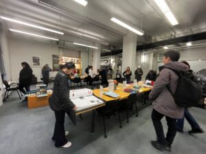 Faculty, students, and staff gathered around a table in a bright, modern library examining archival materials laid out for them on long, rectangular tables covered with various papers, pamphlets, and artists’ books. The people are dressed in casual to semi-formal attire; jackets, jeans or trousers are prevalent. At least one person appears to be wearing headphones. The overall atmosphere is one of focused attention and quiet interaction.The modern space is characterized by high ceilings, white walls, and what seem to be long, linear fluorescent light fixtures. The floor is a light gray carpet. There are visible pillars and some architectural detailing, emphasizing the height of the space. The color palette is subdued, mainly consisting of grays, whites, and muted colors from the clothing and printed materials.