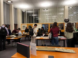 A high-angle view shows a group of people examining archival materials spread out on long tables in a brightly lit, modern room. Some items appear to be in folders and binders and others large books opened up and unfolded maps. The people are mostly seen from the back or sides, their heads bowed over the materials. They are students or researchers, dressed in casual but neat clothing – jeans, sweaters, and jackets with backpacks worn by and next to some individuals. The overall mood is one of calm concentration and scholarly pursuit.