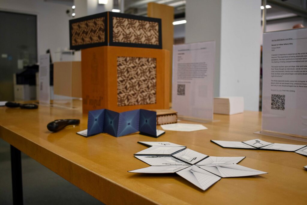 An interactive artists' book display at Books and Bites spring 2025 Showcase on a light wood table featuring various educational materials and artifacts. The centerpiece is a tall, orange and black display artists’ book with woven strips of paper. In front of it is a blue accordion-folded artists’ book, and another part of the same book with a white background arranged in a star shape.