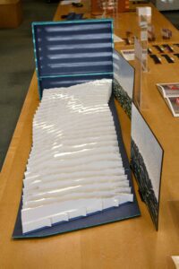 Artists’ book with blue background and varied cut strips of wave-like white paper displayed on a tabletop.