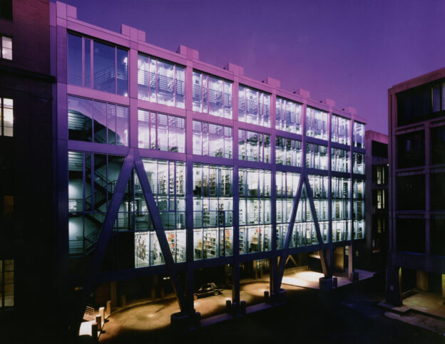 The exterior of Rotch Library at dusk bathed in purple light. Book stacks are visible on multiple floors through the windows.